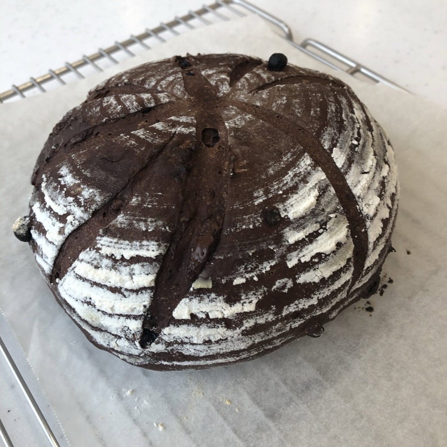 A round cocoa loaf on a cooling rack, dusted with flour and deeply scored in a radiating pattern.